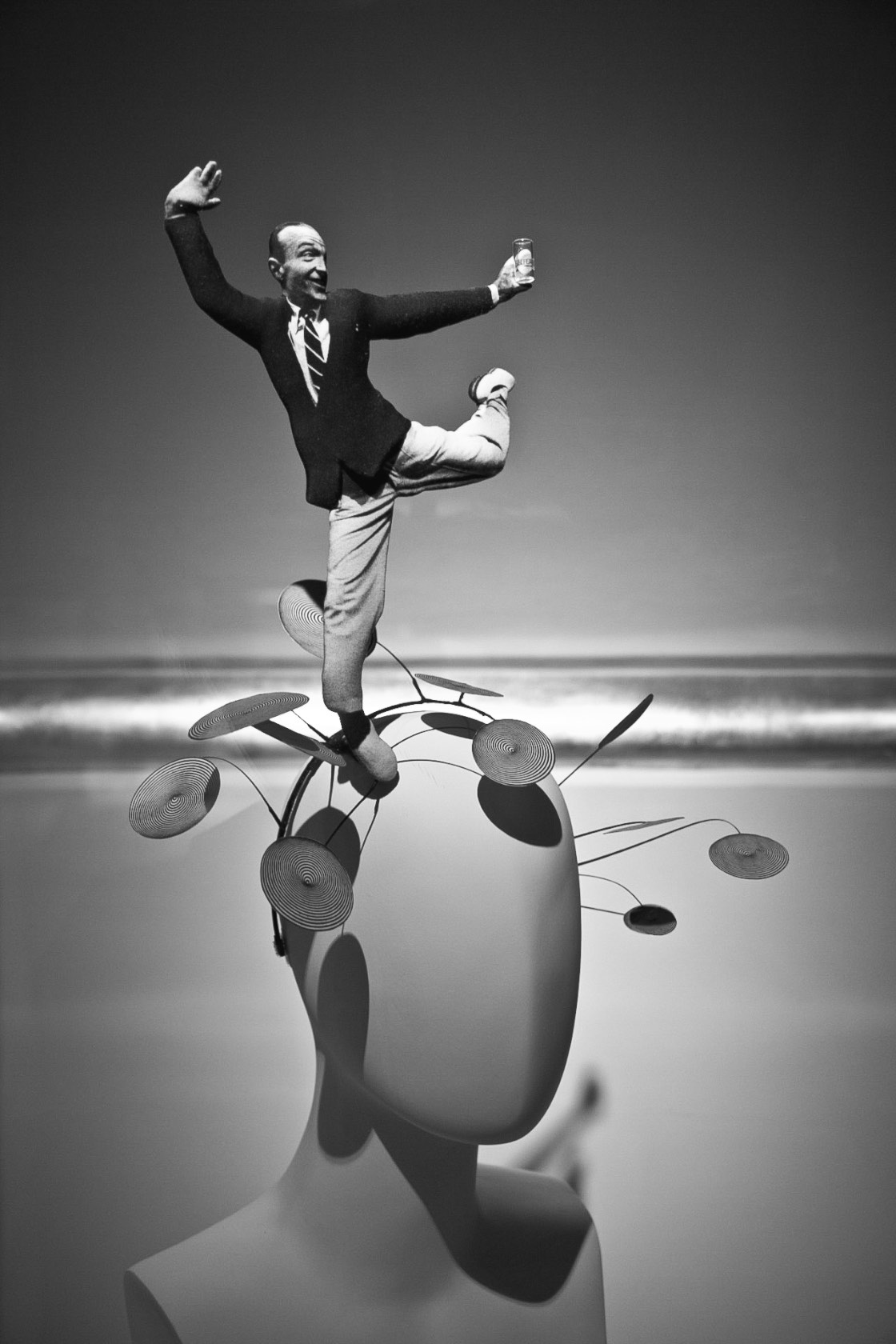 Man in dark suit and striped tie balancing on one leg atop a large rounded hat with spiral metallic disc ornaments extending upward