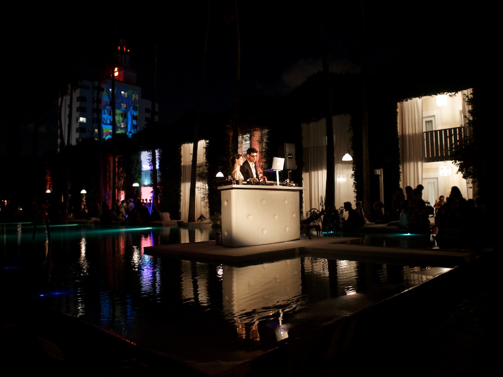 Nighttime photograph of an illuminated circular DJ booth with a figure at the controls, surrounded by lit building facades and reflective water pools.