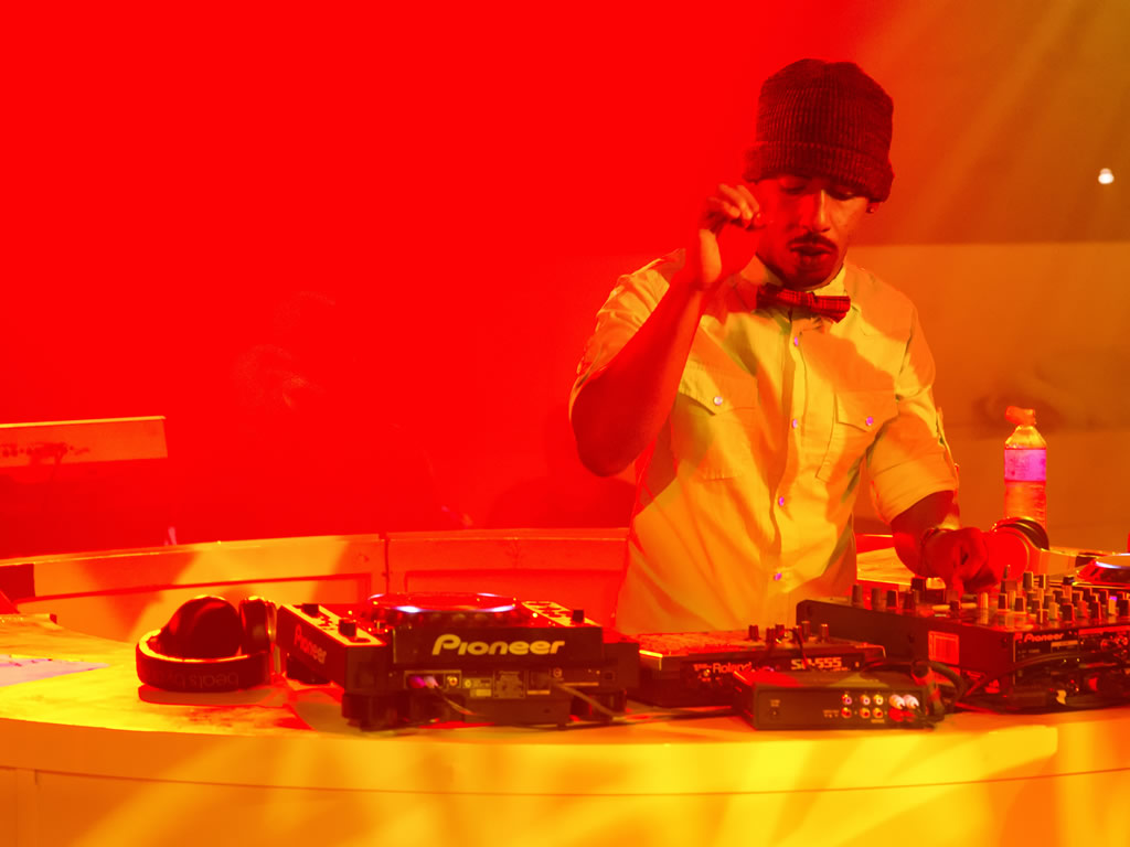 DJ performing at turntables under warm red and orange stage lighting, wearing yellow shirt and dark beanie hat
