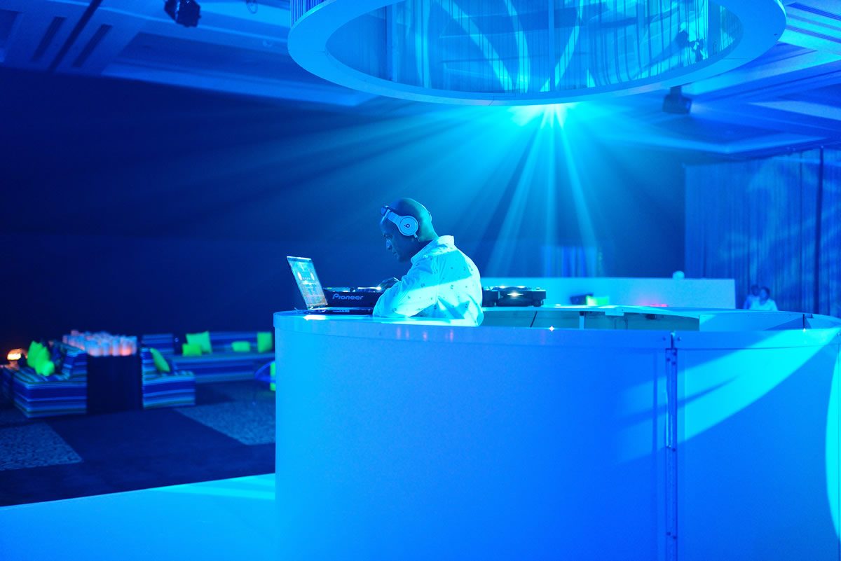 DJ performing at illuminated booth under dramatic blue stage lighting in nightclub setting, wearing headphones