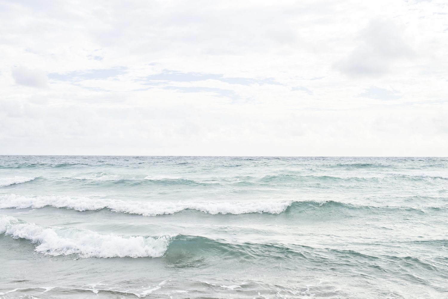 Serene seascape with layered ocean waves rolling toward shore under an overcast sky with soft clouds.