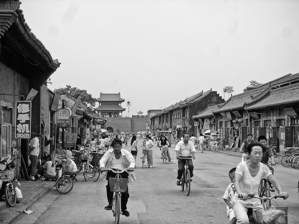 Black and white photograph of a crowded street in ancient Pingyao, China, filled with people riding bicycles and walking past traditional buildings with curved roofs.